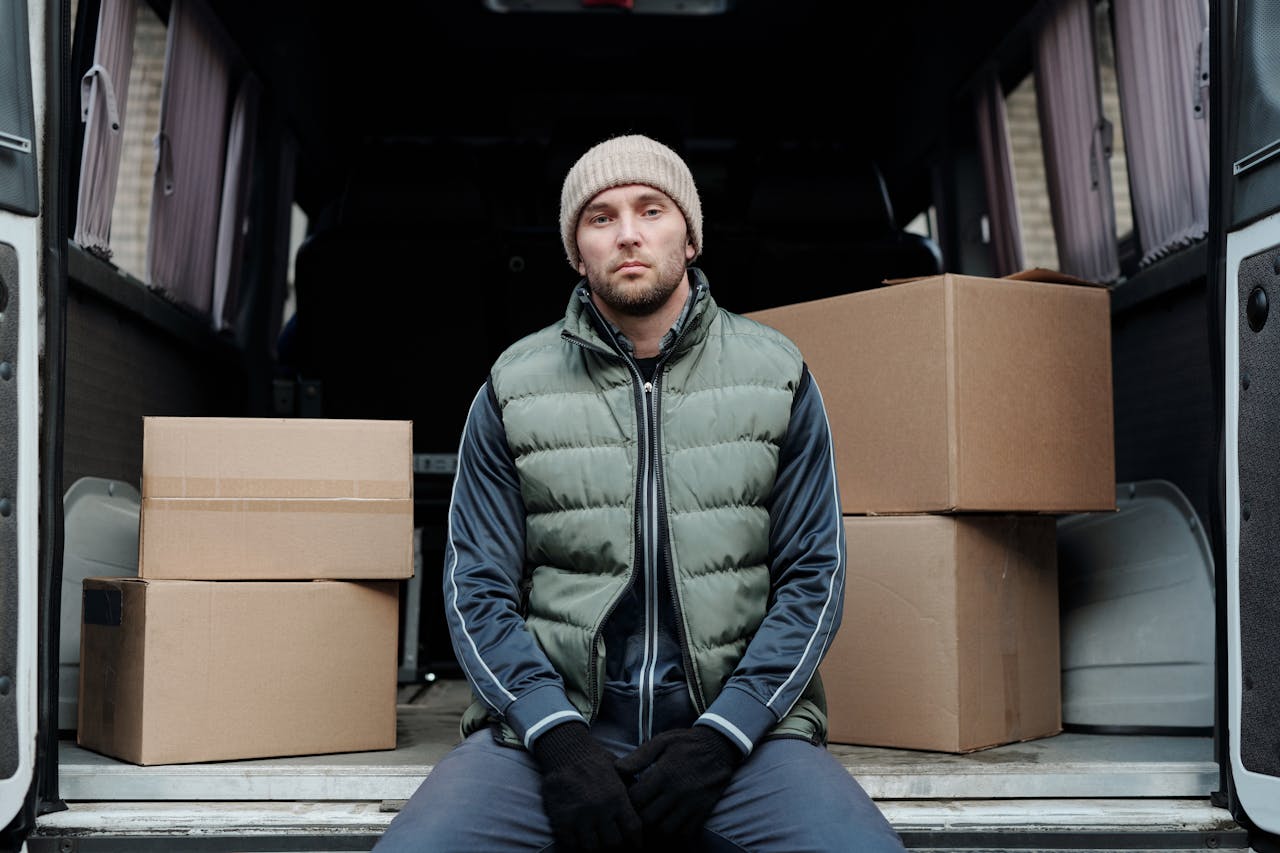 A delivery worker wearing a beanie and vest sits inside a van with cardboard boxes.